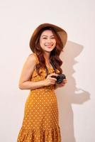 porträtt av glad leende ung kvinna tar Foto med inspiration och bär sommar klänning. flicka innehav retro kamera. modell Framställ på beige bakgrund i hatt