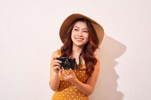 porträtt av glad leende ung kvinna tar Foto med inspiration och bär sommar klänning. flicka innehav retro kamera. modell Framställ på beige bakgrund i hatt