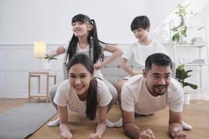 lycklig välbefinnande asiatisk thai familj, barn leker och retar sina föräldrar medan yoga fitnessträning och hälsoträning tillsammans i det vita vardagsrummet, inhemsk livsstil, helgaktivitet. foto