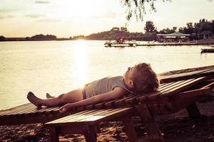 små pojke avkopplande på däck stol på de strand på solnedgång. foto