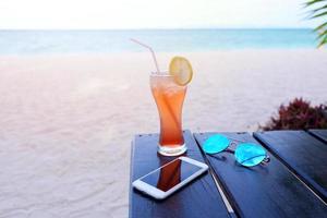 gott färgrik iced cocktail med citron- skiva och solglasögon med smartphone på de tabell i de ö strand, semester begrepp foto