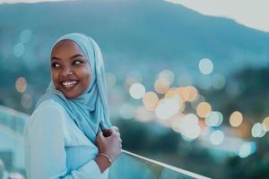 afrikansk muslim kvinna i de natt på en balkong leende på de kamera med stad bokeh lampor i de bakgrund. foto