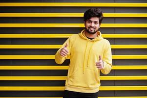 urban ung hipster indisk man i en modern gul tröja. Häftigt söder asiatisk kille ha på sig luvtröja mot randig bakgrund som visar tumme upp. foto