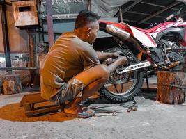 ett gammal man i kort shorts är påfrestande hård till förändra de bak- motorcykel däck på natt i hans verkstad. Foto tagen på natt utomhus i luhu by, gorontalo på augusti 21, 2022