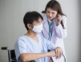 asiatisk kvinna läkare använder ett stetoskop för att kontrollera lungrytmen hos en manlig patient som bär ansiktsmask medan han sitter i en rullstol på sjukhuset. foto