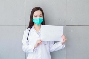asiatisk kvinna läkare bär medicinsk täcka och ansikte mask medan visar enkel papper vit papper till närvarande något i hälsa skydd begrepp. foto