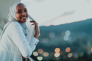 afrikansk muslim kvinna i de natt på en balkong leende på de kamera med stad bokeh lampor i de bakgrund. foto