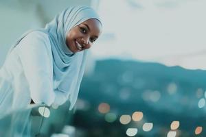 afrikansk muslim kvinna i de natt på en balkong leende på de kamera med stad bokeh lampor i de bakgrund. foto