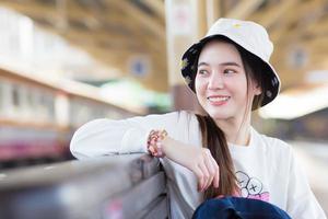 asiatisk skön flicka i en långärmad vit skjorta och en hatt sitter Lycklig smilie i de tåg station väntar för de tåg till anlända. foto
