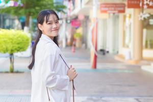 självsäker ung företag asiatisk arbetssätt kvinna vem bär en vit skjorta och axel väska ler lyckligt medan hon är gående till arbete på kontor i de stad med en affär byggnad i de bakgrund. foto