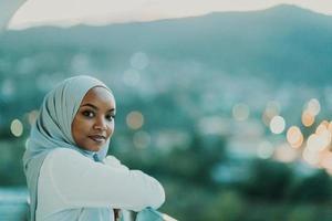 afrikansk muslim kvinna i de natt på en balkong leende på de kamera med stad bokeh lampor i de bakgrund. foto