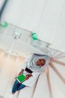 ung afrikansk modern muslim kvinna använder sig av läsplatta dator medan Sammanträde på de trappa på Hem bär hijab kläder topp se foto