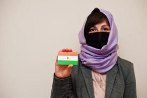 porträtt av ung muslim kvinna bär formell ha på sig, skydda ansikte mask och hijab huvud scarf, håll niger flagga kort mot isolerat bakgrund. coronavirus Land begrepp. foto