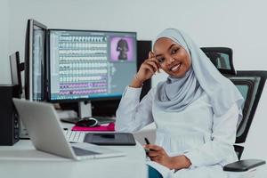 ung Afroamerikan modern muslim affärskvinna bär en scarf i en kreativ ljus kontor arbetsplats med en stor skärm. foto