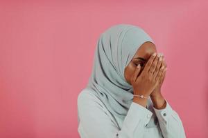 modern afrikansk muslim kvinna gör traditionell bön till Gud, håller händer i bön- gest, bär traditionell vit kläder, har allvarlig ansiktsbehandling uttryck, isolerat över plast rosa bakgrund foto