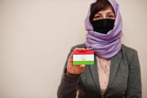 porträtt av ung muslim kvinna bär formell ha på sig, skydda ansikte mask och hijab huvud scarf, håll tadzjikistan flagga kort mot isolerat bakgrund. coronavirus Land begrepp. foto