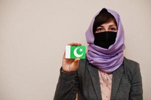 porträtt av ung muslim kvinna bär formell ha på sig, skydda ansikte mask och hijab huvud scarf, håll pakistan flagga kort mot isolerat bakgrund. coronavirus Land begrepp. foto
