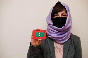 porträtt av ung muslim kvinna bär formell ha på sig, skydda ansikte mask och hijab huvud scarf, håll maldiverna flagga kort mot isolerat bakgrund. coronavirus Land begrepp. foto