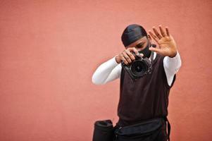 ung professionell afrikansk amerikan videographer innehav professionell kamera med proffs Utrustning. afro kameraman bär svart duraq och ansikte skydda mask, framställning en videoklipp. foto