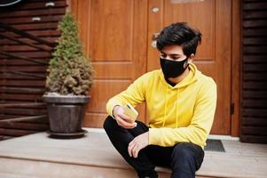 urban ung hipster indisk man i en modern gul tröja med mobil telefon. Häftigt söder asiatisk kille ha på sig luvtröja och svart ansikte skydda mask, under ny vanligt. foto