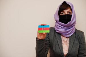 porträtt av ung muslim kvinna bär formell ha på sig, skydda ansikte mask och hijab huvud scarf, håll azerbaijan flagga kort mot isolerat bakgrund. coronavirus Land begrepp. foto