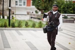 ung professionell afrikansk amerikan videographer innehav professionell kamera med proffs Utrustning. afro kameraman bär svart duraq framställning en videoklipp. foto