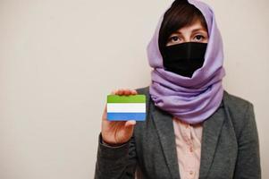 porträtt av ung muslim kvinna bär formell ha på sig, skydda ansikte mask och hijab huvud scarf, håll sierra leone flagga kort mot isolerat bakgrund. coronavirus Land begrepp. foto