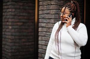 glamorös afrikansk amerikan kvinna i vit turtleneck Tröja utgör på gata med mobil telefon. foto