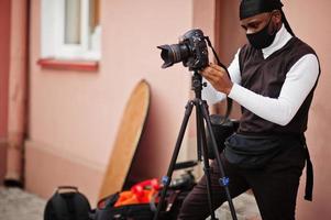 ung professionell afrikansk amerikan videographer innehav professionell kamera med proffs Utrustning. afro kameraman bär svart duraq och ansikte skydda mask, framställning en videoklipp. foto