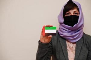 porträtt av ung muslim kvinna bär formell ha på sig, skydda ansikte mask och hijab huvud scarf, håll förenad arab emirates flagga kort mot isolerat bakgrund. coronavirus Land begrepp. foto
