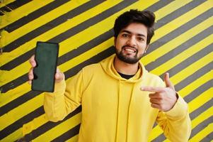 urban ung hipster indisk man i en modern gul tröja. Häftigt söder asiatisk kille ha på sig luvtröja mot randig bakgrund visa finger till skärm av mobil telefon. foto