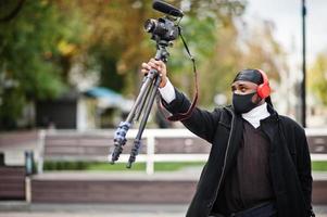 ung professionell afrikansk amerikan videographer innehav professionell kamera med proffs Utrustning. afro kameraman bär svart duraq och ansikte skydda mask, framställning en videoklipp. foto