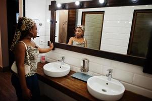 härlig afrikansk amerikan kvinna med dreadlocks i leopard utrusta ser i de spegel på toalett Kafé. skön Häftigt modern svart ung flicka inomhus. foto