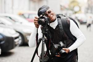 ung professionell afrikansk amerikan videographer innehav professionell kamera med stativ proffs Utrustning. afro kameraman bär svart duraq framställning en videoklipp. foto
