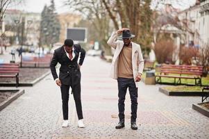 två mode svart män gående på gata. modern porträtt av afrikansk amerikan manlig modeller. ha på sig kostym, täcka och hatt. foto