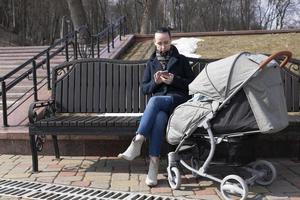 ung kvinna promenader i de parkera i de vår med en bebis transport. mor utseende in i en mobil telefon medan de bebis sover utomhus. foto