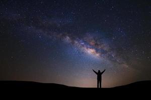 landskap med mjölkig sätt galax och Plats damm i de universum, natt himmel med stjärnor och silhuett av en stående sportig man med Uppfostrad upp vapen på hög berg. foto