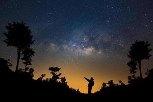 en man är stående Nästa till de mjölkig sätt galax pekande på en ljus stjärna i skog, lång exponering fotografera, med spannmål. foto
