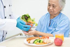 asiatisk senior eller äldre gammal dam kvinna patienten äter frukost och grönsaker hälsosam mat med hopp och glad medan du sitter och hungrig på sängen på sjukhuset. foto