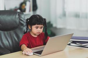 asiatisk bebis flicka bär en röd t-shirt använda sig av bärbar dator och studie uppkopplad på trä tabell skrivbord i vardagsrum på Hem. utbildning inlärning uppkopplad från Hem begrepp. foto