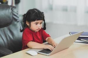 asiatisk bebis flicka bär en röd t-shirt använda sig av bärbar dator och studie uppkopplad på trä tabell skrivbord i vardagsrum på Hem. utbildning inlärning uppkopplad från Hem begrepp. foto