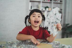 asiatisk bebis flicka bär en röd t-shirt innehav dollar räkningen på trä tabell skrivbord i vardagsrum på Hem. sparande investering rikedom begrepp. foto