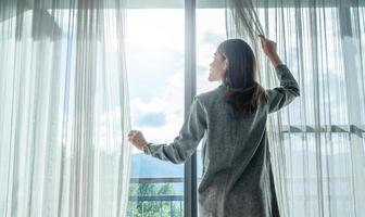 bak- se av lycka asiatisk kvinna öppen gardiner för ser skön se utanför henne rum. foto