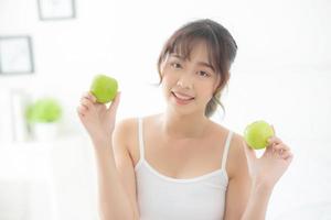 vackert porträtt ung asiatisk kvinna som håller och äter grönt äpple frukt i sovrummet hemma, livsstil för näring flicka hälsosam och omsorg viktminskning, hälsa och välbefinnande koncept. foto