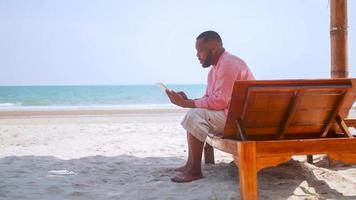 affärsman spenderar hans helger avkopplande på de strand. mogna afrikansk amerikan man använder sig av en läsplatta medan Sammanträde på de strand. foto