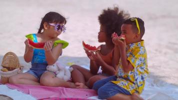 grupp av barn äter vattenmeloner medan har en picknick på de strand under sommar semester. foto