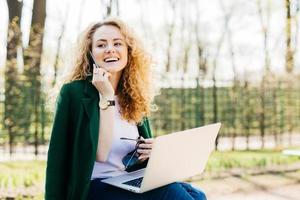 ung kvinnlig entreprenör med fluffigt hår som sitter utanför framför öppen generisk bärbar dator och pratar i mobiltelefon med glad blick håller solglasögon i handen med bärbar dator och tittar åt sidan foto