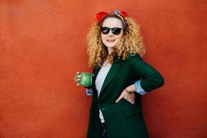 trendig ung vacker kvinna med fluffigt blont hår bär pannband, solglasögon och jacka som håller i ena handen en kopp varmt kaffe och andra handen på hennes midja poserar i studion mot den orange väggen foto