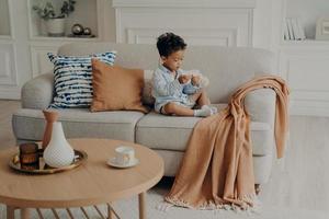 litet härligt afroamerikanskt barn som sitter på soffan och leker ensam i vardagsrummet foto