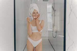 ung kvinna med vältränad kropp i vita klassiska underkläder som står i badrummet och borstar tänderna foto